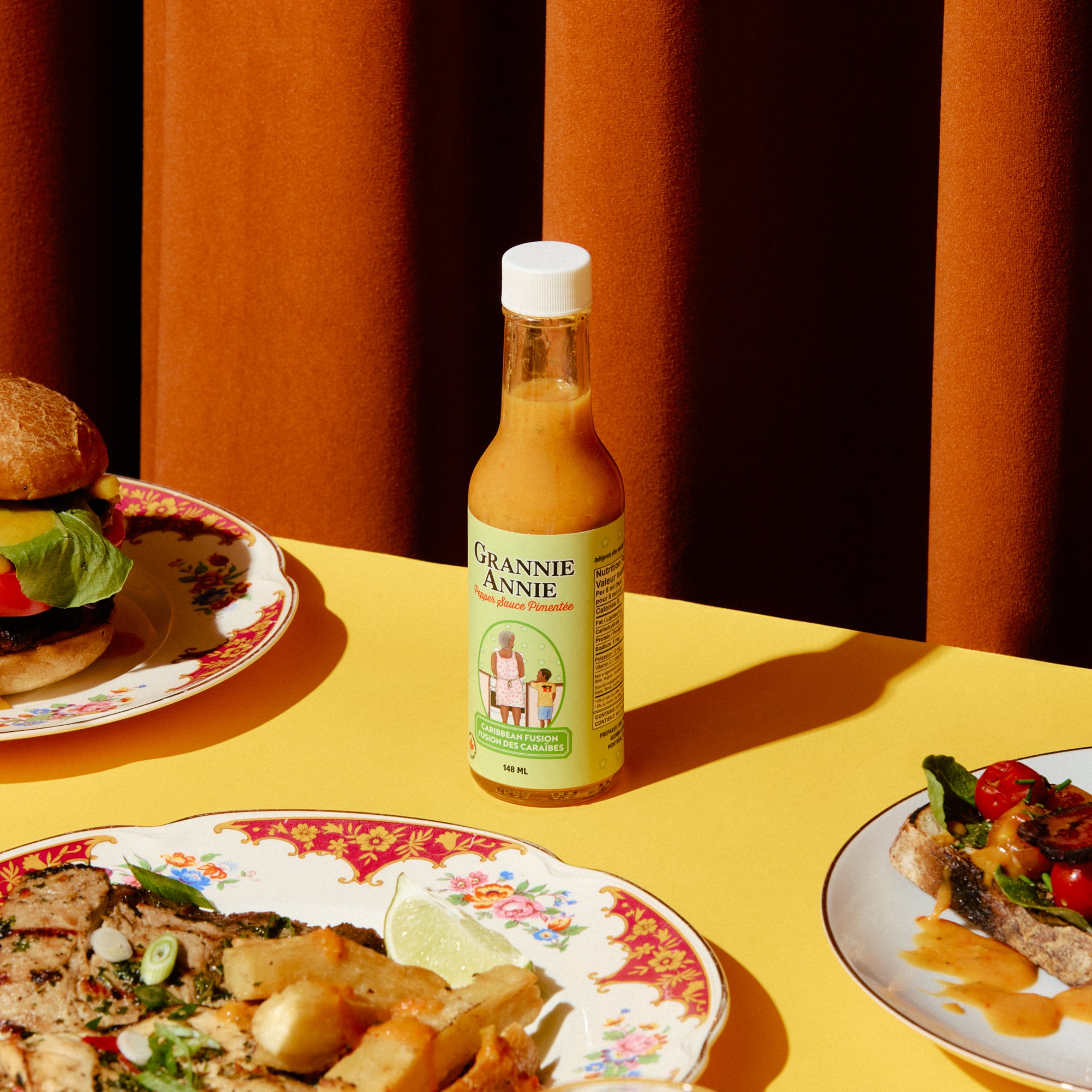 Bottle of pepper sauce on a table with plates of food and a glass of red liquid.
