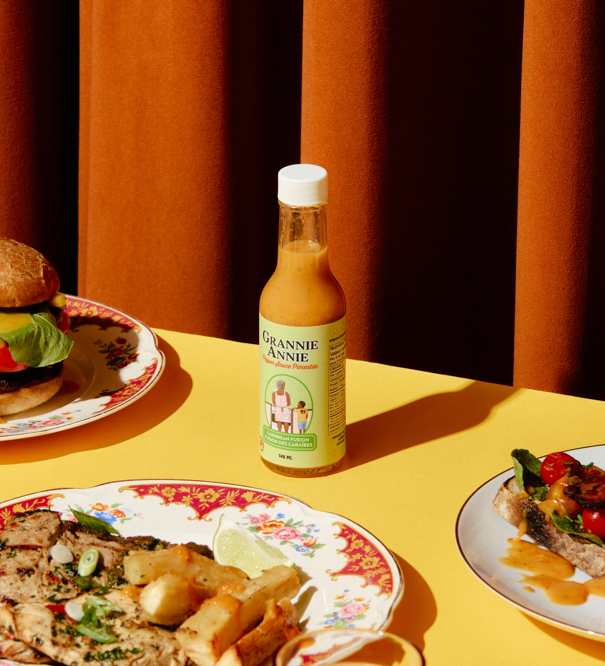 Bottle of pepper sauce on a table with plates of food and a glass of red liquid.