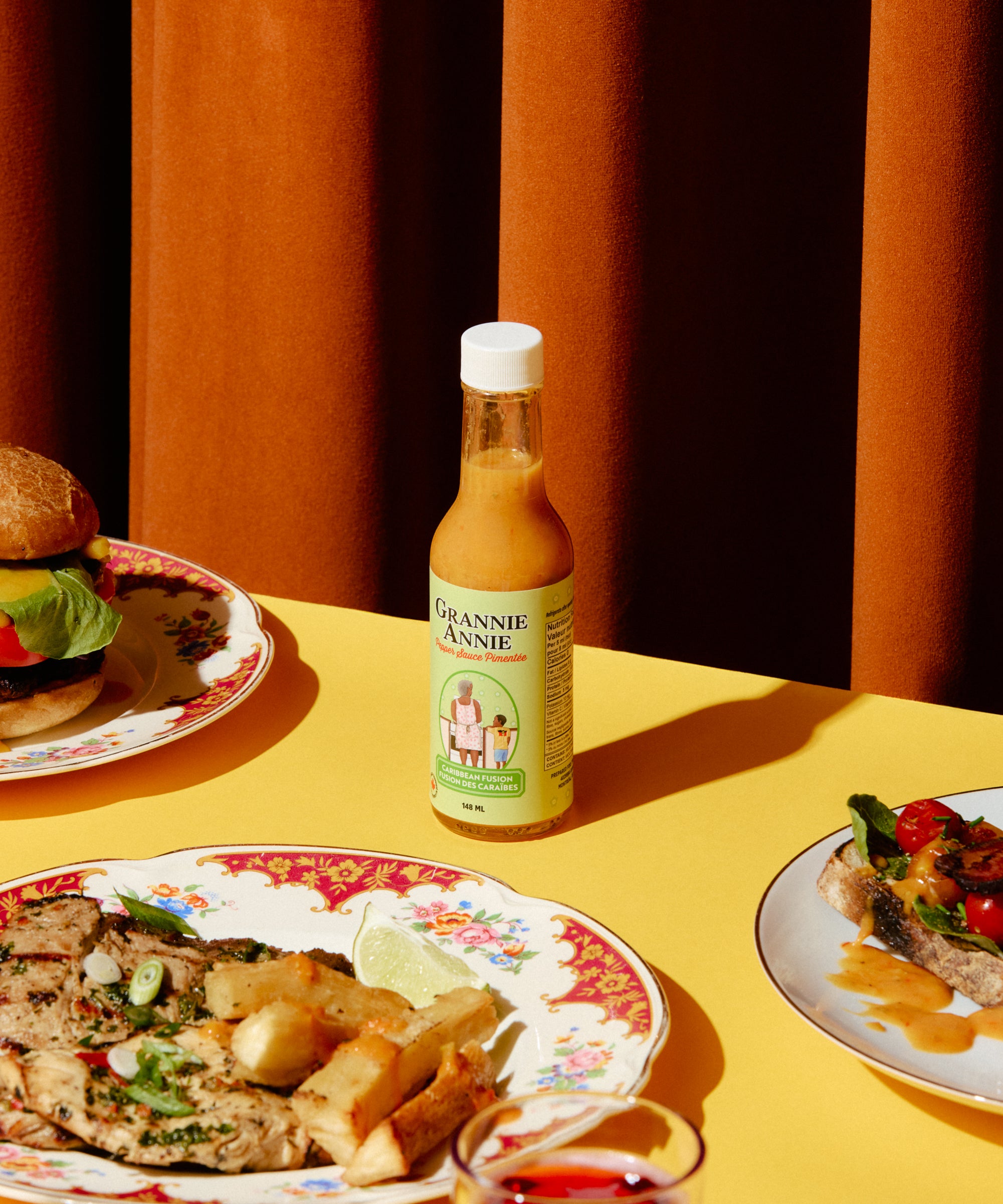 Bottle of pepper sauce on a table with plates of food and a glass of red liquid.